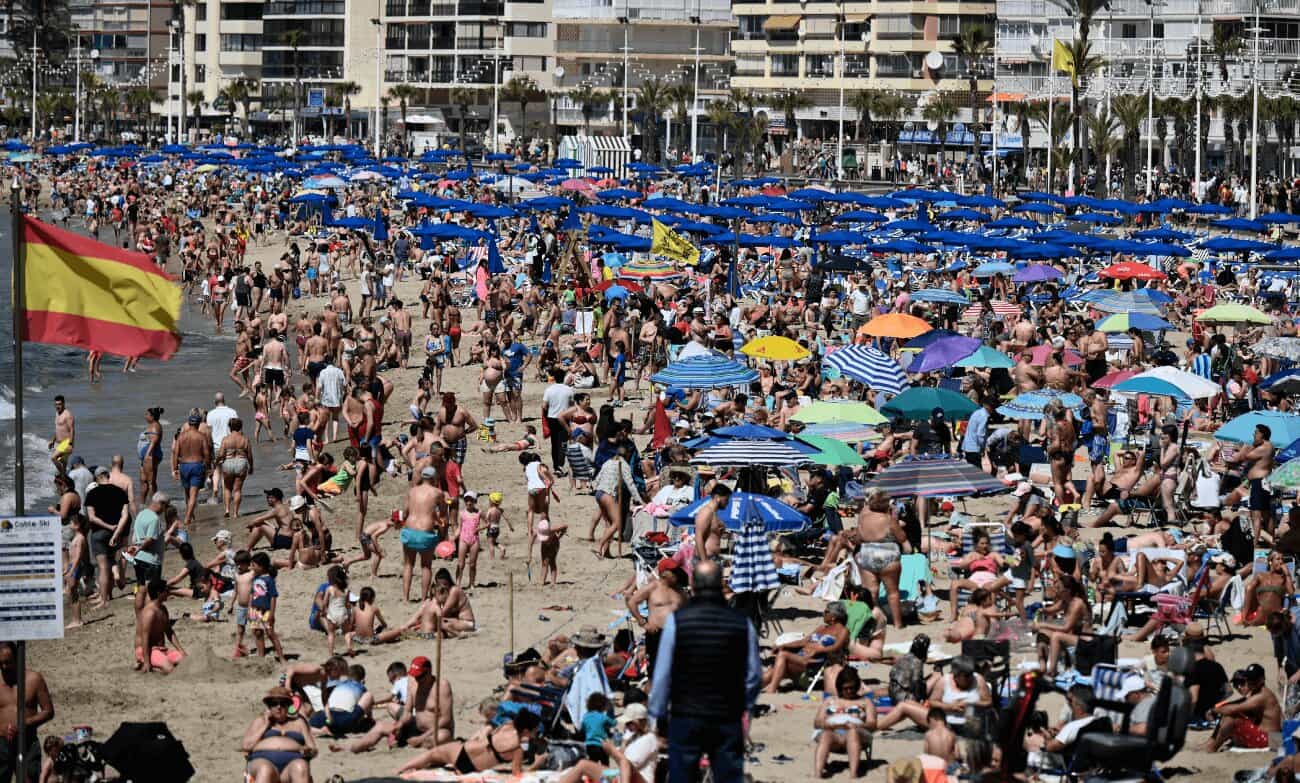 La playa de Benidorm completamente abarrotada de bañistas y sombrillas, con una bandera de España en primer plano.